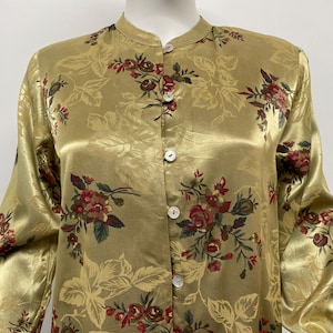 May include: A golden-yellow, long-sleeved blouse with a mandarin collar and a floral pattern in burgundy and green. The blouse has a button-up front with white buttons and a silky sheen.