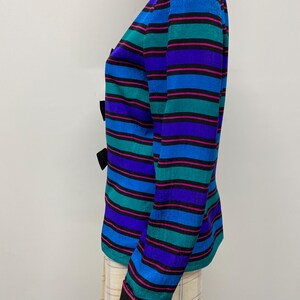 May include: A colorful striped blazer with black bows. The blazer features horizontal stripes in shades of blue, teal, purple, and pink. The sleeves have black trim. The blazer is displayed on a mannequin.