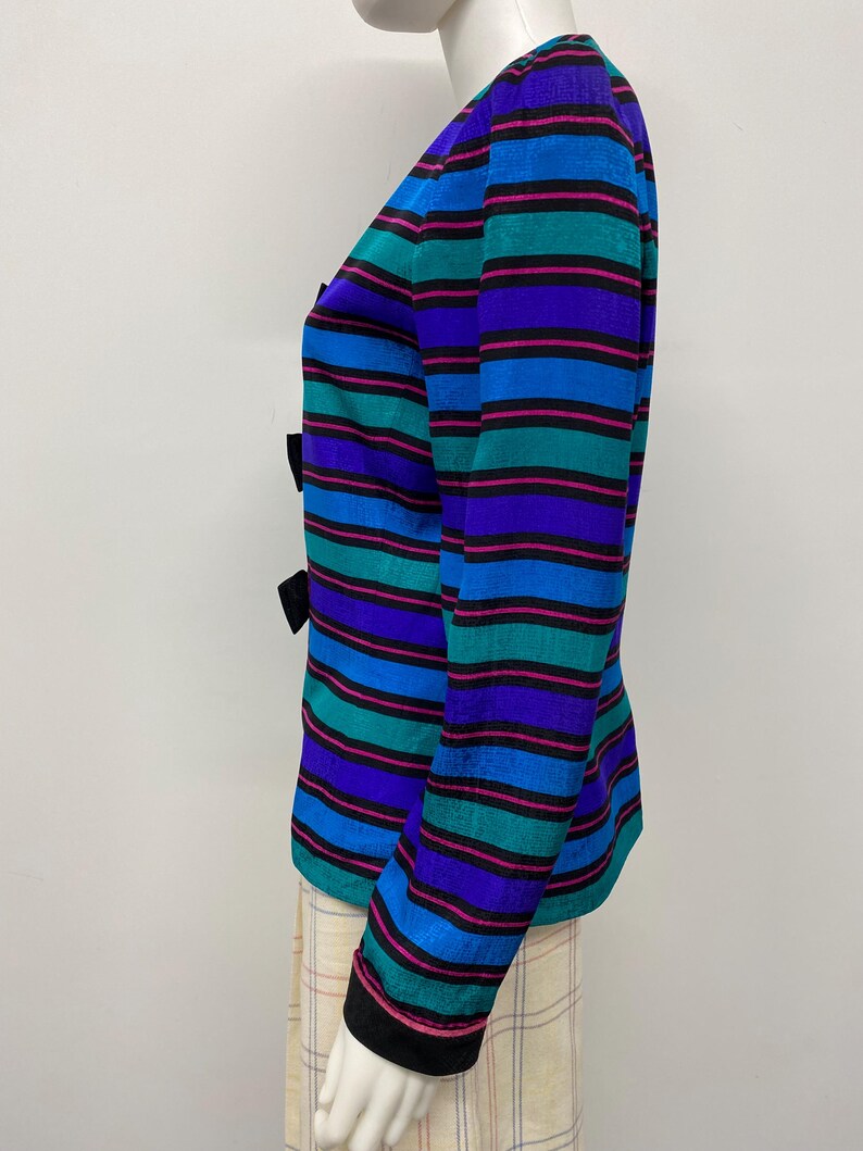 May include: A colorful striped blazer with black bows. The blazer features horizontal stripes in shades of blue, teal, purple, and pink. The sleeves have black trim. The blazer is displayed on a mannequin.