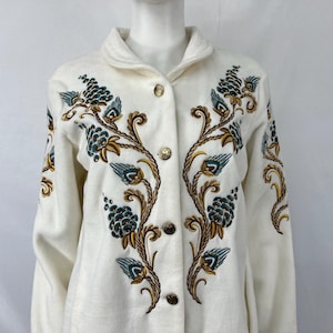 May include: Off-white coat with floral embroidery. The coat features a collar, gold-colored buttons, and detailed embroidery in shades of teal, brown, and gold. The floral design is symmetrical, with vines and clusters of leaves.