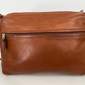 May include: A brown leather crossbody bag with a rectangular shape. The bag features a zippered front pocket, a long adjustable strap, and gold-toned hardware. The leather has a smooth texture and a slight sheen.