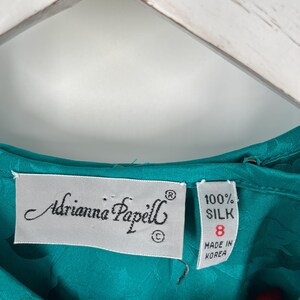 May include: Close-up of a teal silk garment with the label "Adrianna Papell." The label indicates the garment is size 8 and made of 100% silk. The fabric has a subtle floral pattern.