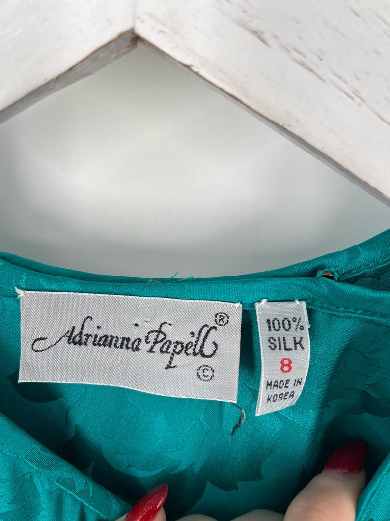 May include: Close-up of a teal silk garment with the label "Adrianna Papell." The label indicates the garment is size 8 and made of 100% silk. The fabric has a subtle floral pattern.