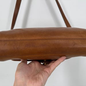 May include: A brown leather shoulder bag with a long strap. The bag has a rectangular shape with rounded edges and a brass clasp. The leather shows signs of wear, adding to its vintage appeal.