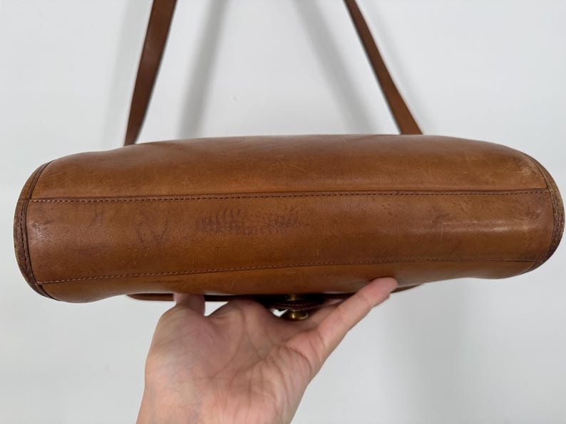 May include: A brown leather shoulder bag with a long strap. The bag has a rectangular shape with rounded edges and a brass clasp. The leather shows signs of wear, adding to its vintage appeal.