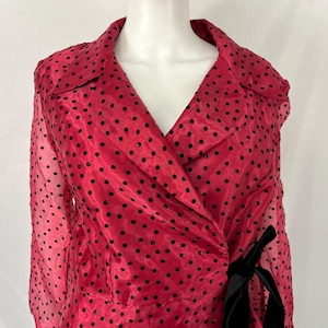 May include: A red, sheer wrap top with black polka dots. The top features a notched collar, long sleeves, and a black velvet tie at the waist. The fabric has a slight sheen.