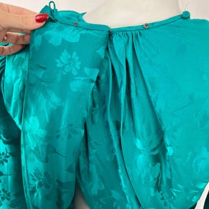May include: Teal-colored dress with a floral pattern. The dress features a high neckline with small silver-colored buttons and a keyhole back. The fabric has a shiny, satin-like appearance. The sleeves are long and gathered at the shoulders.