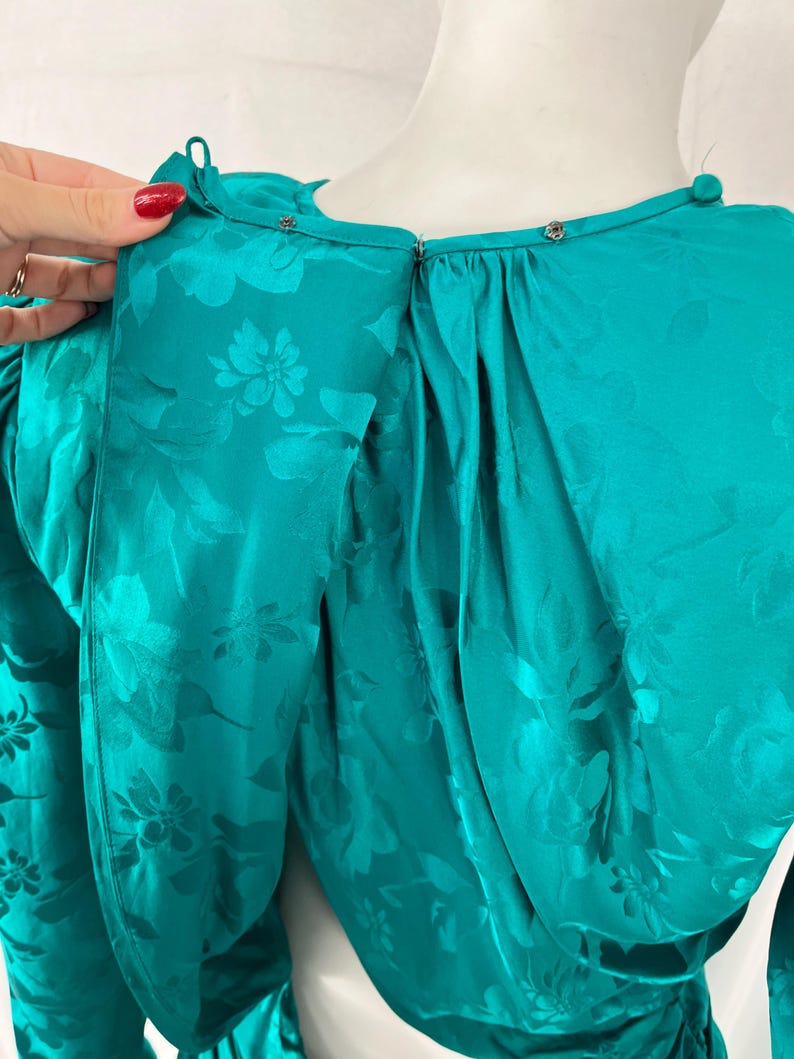 May include: Teal-colored dress with a floral pattern. The dress features a high neckline with small silver-colored buttons and a keyhole back. The fabric has a shiny, satin-like appearance. The sleeves are long and gathered at the shoulders.