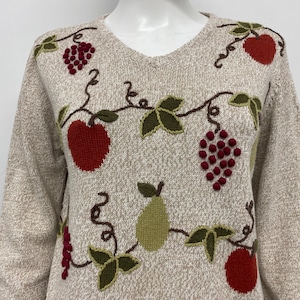 May include: A beige long-sleeve sweater featuring an embroidered fruit pattern. The design includes red apples, green pears, and clusters of dark red grapes, all connected by brown vine stems and dark green leaves across the front of the garment.