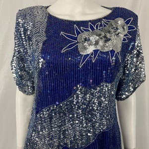 May include: A blue sequined dress with silver accents. The dress features a round neckline and short sleeves. A floral design with silver sequins is on the upper chest. The dress is made of a shiny, reflective material.