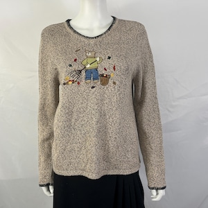 May include: A long-sleeved, speckled beige knit sweater featuring a crew neckline with dark trim. The center of the chest displays an embroidered graphic of a teddy bear wearing a green shirt and blue jeans, holding a rake near a basket of leaves.