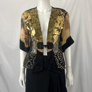 May include: A sheer, open-front jacket with a floral pattern in gold, brown, and black. The jacket features black lace detailing and a decorative closure. The sleeves are short, and the garment is displayed on a mannequin.