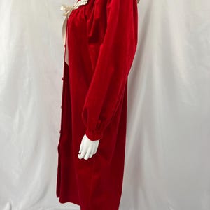 Op de afbeelding: Een lange, rode fluwelen robe met een witte kanten kraag en manchetten. De robe heeft lange mouwen, een knoopsluiting aan de voorkant en een cr&egrave;mekleurig lint. De robe wordt op een mannequin getoond.