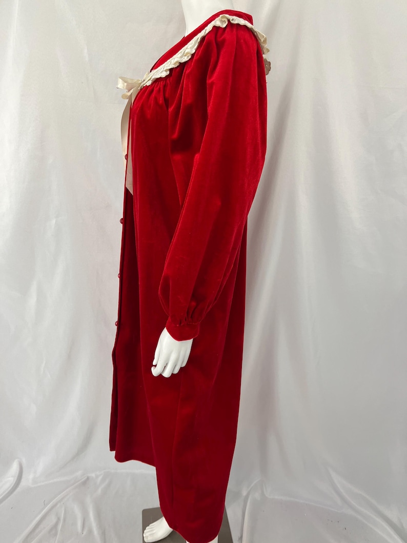 Op de afbeelding: Een lange, rode fluwelen robe met een witte kanten kraag en manchetten. De robe heeft lange mouwen, een knoopsluiting aan de voorkant en een cr&egrave;mekleurig lint. De robe wordt op een mannequin getoond.