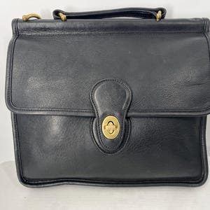May include: Black leather briefcase with a top handle and a flap closure. The briefcase features a gold-tone turn-lock clasp and hardware. The bag has a structured design and appears to be vintage.