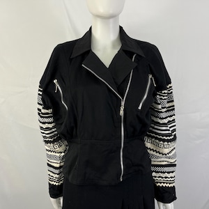 May include: A black jacket featuring a diagonal silver zipper closure, a notched collar, and two slanted zippered pockets on the chest. The long sleeves are decorated with a contrasting white and black geometric pattern and textured trim.