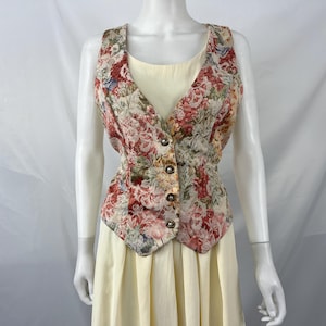 May include: A sleeveless vest featuring a multi-colored floral pattern in shades of red, pink, and green, secured with four metallic buttons down the front, worn over a solid pale yellow dress with a scoop neckline and a pleated skirt.