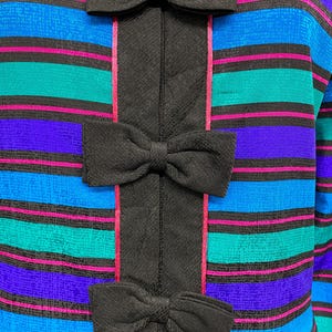 May include: Close-up of a colorful top with horizontal stripes in teal, blue, purple, and pink. The top features a black vertical panel down the center with three black bow accents.