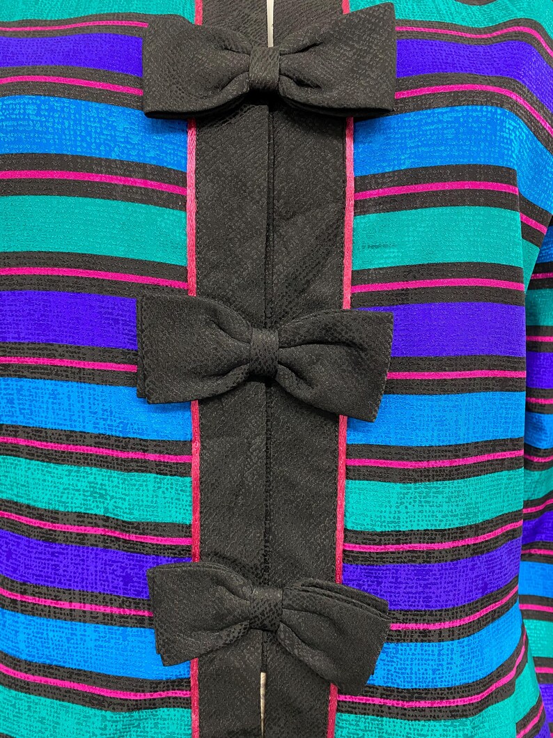 May include: Close-up of a colorful top with horizontal stripes in teal, blue, purple, and pink. The top features a black vertical panel down the center with three black bow accents.
