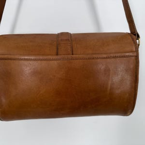 May include: A brown leather crossbody bag with a rectangular shape. The bag features a flap closure with a buckle and a long adjustable strap. The leather has a worn, vintage appearance, with visible grain and slight variations in color.