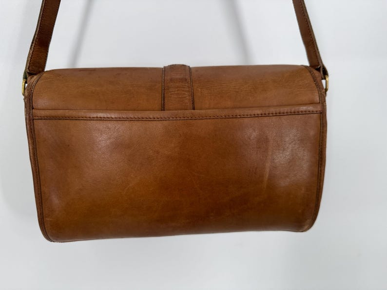 May include: A brown leather crossbody bag with a rectangular shape. The bag features a flap closure with a buckle and a long adjustable strap. The leather has a worn, vintage appearance, with visible grain and slight variations in color.