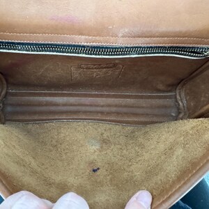 May include: Open view of a vintage brown leather Coach handbag. The interior features a suede-like lining and a zippered pocket. The bag shows signs of wear, with visible scuffs and marks, adding to its character. The Coach logo is visible.