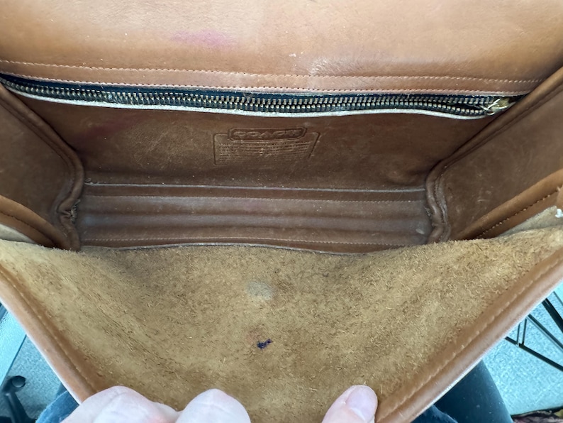 May include: Open view of a vintage brown leather Coach handbag. The interior features a suede-like lining and a zippered pocket. The bag shows signs of wear, with visible scuffs and marks, adding to its character. The Coach logo is visible.