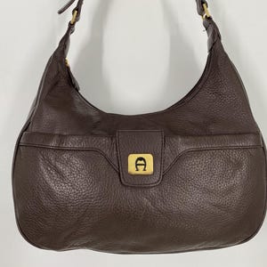 May include: A brown leather shoulder bag with a pebbled texture. The bag has a curved shape, a front pocket, and a flap closure with a gold-colored logo. The strap is also brown leather.