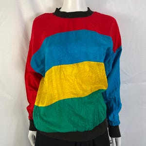 May include: A long-sleeve pullover shirt featuring a color-blocked design with horizontal sections of red, blue, yellow, and green. The garment has a solid black crew neckline, black sleeve cuffs, and a black hemline, constructed from a smooth fabric.