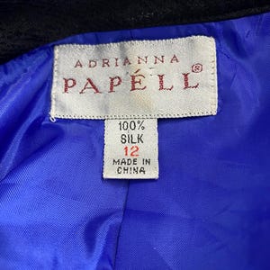 May include: Close-up of a label from an Adrianna Papell garment. The label is rectangular with the brand name in burgundy. The label also indicates the garment is 100% silk, size 12, and made in China. The lining is bright blue.