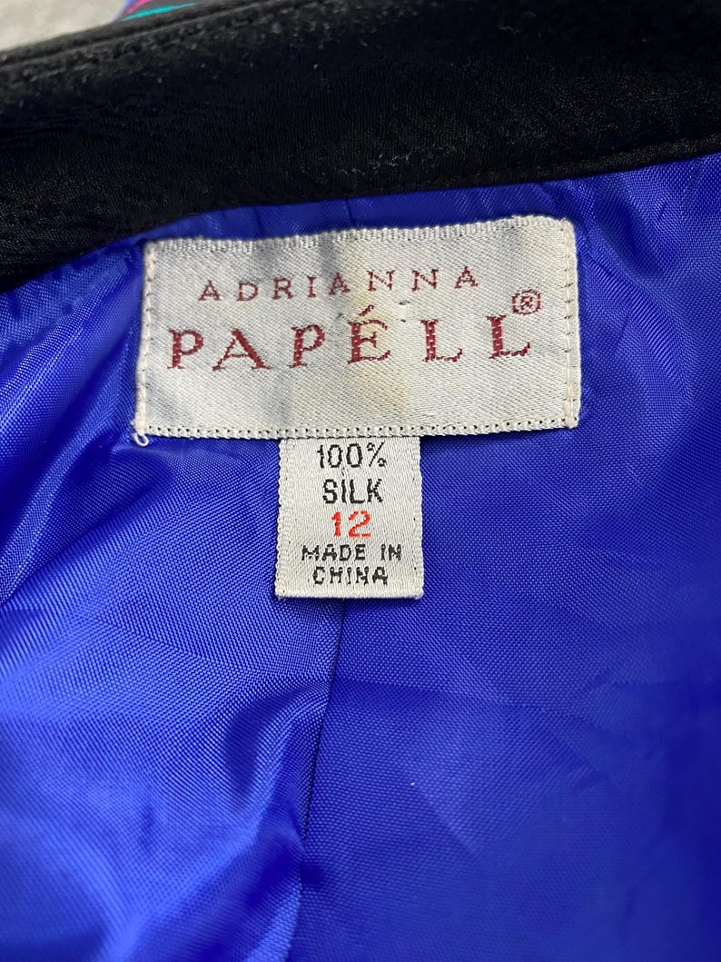 May include: Close-up of a label from an Adrianna Papell garment. The label is rectangular with the brand name in burgundy. The label also indicates the garment is 100% silk, size 12, and made in China. The lining is bright blue.