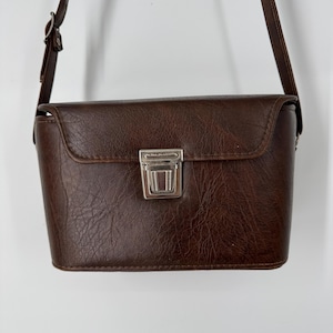 May include: A dark brown rectangular shoulder bag with a textured surface, featuring a silver-toned metal push-lock closure on the front flap and an adjustable thin shoulder strap attached to the sides of the bag.