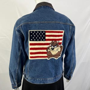 May include: A blue denim jacket featuring a large rectangular patch on the back. The patch displays the American flag with a brown, furry-textured cartoon character of the Tasmanian Devil positioned over the lower right corner of the flag.