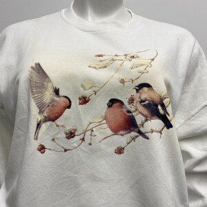 May include: White sweatshirt featuring a detailed illustration of three birds perched on branches with berries. The birds have red, brown, and black plumage. The branches are adorned with berries and leaves.