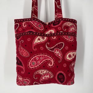May include: A quilted tote bag in a deep red paisley pattern. The bag features a repeating design of large paisley shapes in shades of red, cream, and black. The bag has two matching red straps.