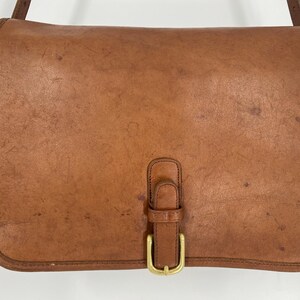 May include: A rectangular tan leather shoulder bag featuring a smooth flap closure secured by a single vertical leather strap and a brass-toned metal buckle. The leather surface displays natural variations in color and texture throughout the exterior.