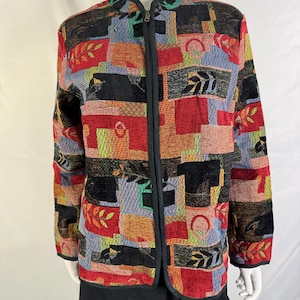 May include: A long-sleeve zip-up jacket featuring a multi-colored tapestry pattern with blocks of red, black, blue, and yellow. The design includes woven leaf motifs and geometric shapes throughout the fabric, finished with a black trim along the collar.