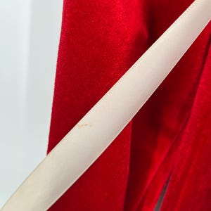 Op de afbeelding: Close-up van een levendige rode fluwelen stof met een cr&egrave;mekleurig lint diagonaal over het frame. Het lint heeft een lichte glans, die contrasteert met de matte textuur van het fluweel. De afbeelding benadrukt de texturen en kleuren.