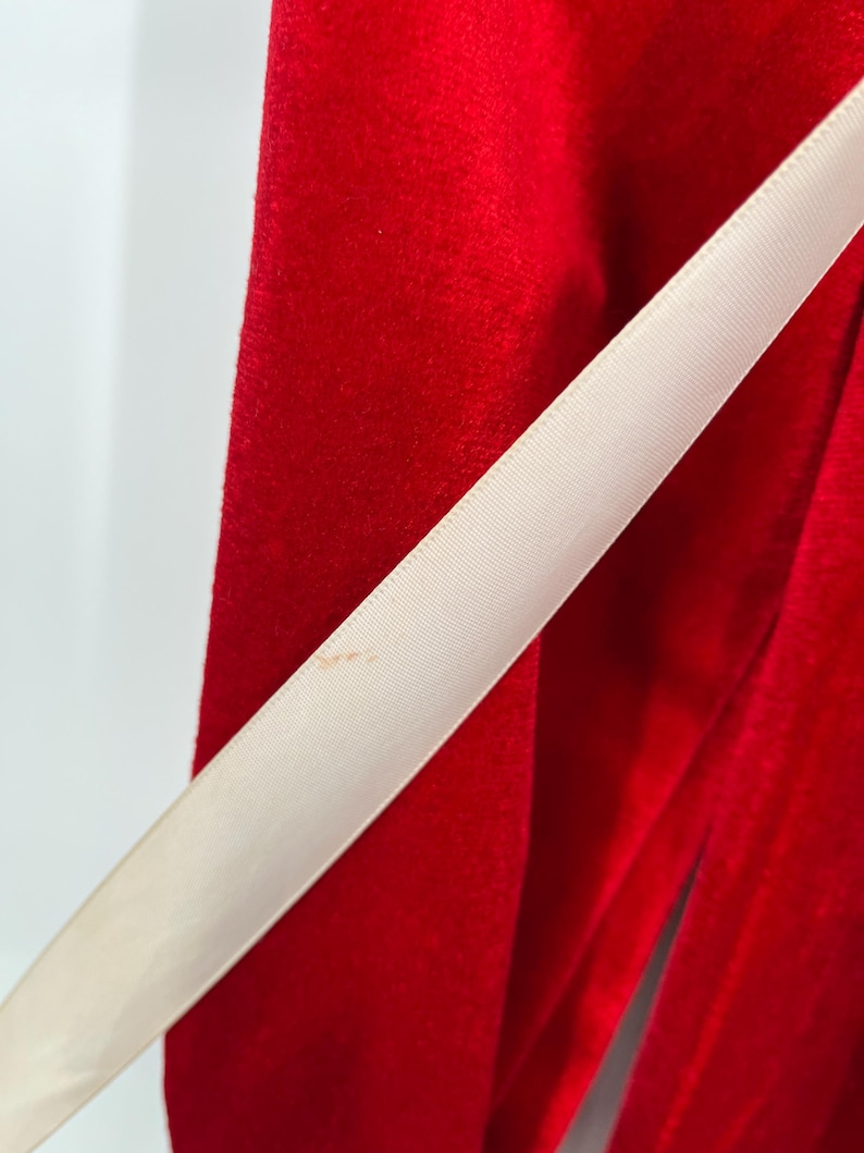 Op de afbeelding: Close-up van een levendige rode fluwelen stof met een cr&egrave;mekleurig lint diagonaal over het frame. Het lint heeft een lichte glans, die contrasteert met de matte textuur van het fluweel. De afbeelding benadrukt de texturen en kleuren.