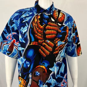 May include: Short-sleeved button-down shirt featuring a vibrant graphic of a superhero in shades of blue, orange, and black. The shirt's background is a pattern of blue shapes. The shirt is displayed on a white mannequin.
