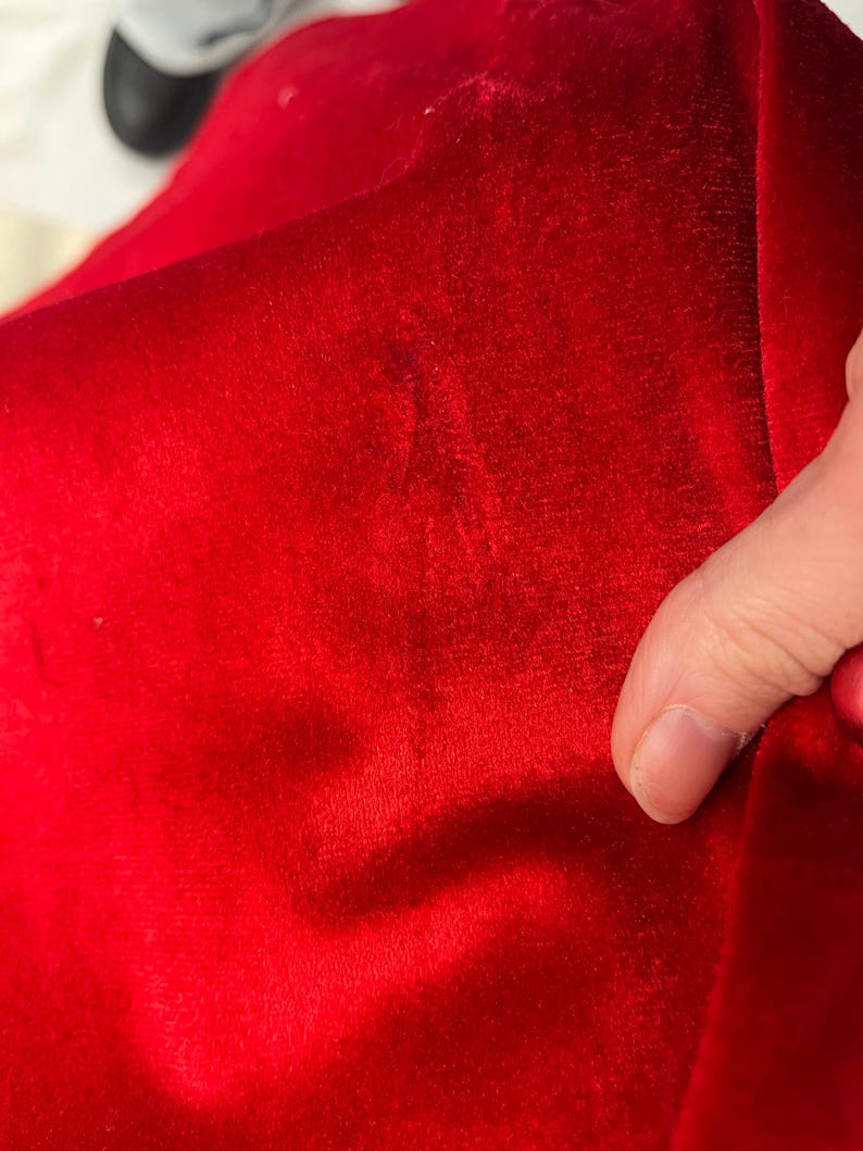 Op de afbeelding: Close-up van een levendige rode fluwelen stof. Het zachte materiaal heeft een zachte textuur en een rijke kleur, met subtiele kleurschakeringen. De stof wordt vastgehouden en toont zijn drape en glans.