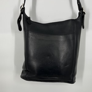 May include: A black leather shoulder bag with a rectangular shape and a front pocket. The bag has a long adjustable strap and silver-tone hardware. The bag is approximately 12 inches wide and 11 inches tall.