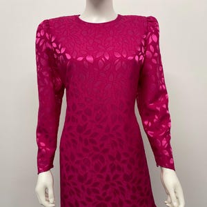May include: A magenta-colored dress with long sleeves and a round neckline. The dress features a repeating leaf pattern across the fabric. The dress is displayed on a mannequin.