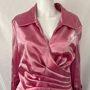 May include: A pink, long-sleeved blouse with a collared neckline and a wrap-style front. The blouse has a shiny, satin-like texture and is displayed on a mannequin. The sleeves have buttoned cuffs.