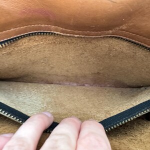 May include: Open view of the interior of a brown leather bag. The bag's interior is a lighter tan color, with a black zipper along the top edge. The leather shows signs of wear and use, with some discoloration.