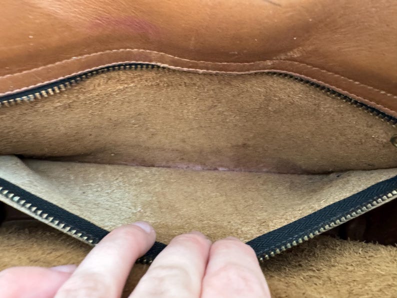 May include: Open view of the interior of a brown leather bag. The bag's interior is a lighter tan color, with a black zipper along the top edge. The leather shows signs of wear and use, with some discoloration.