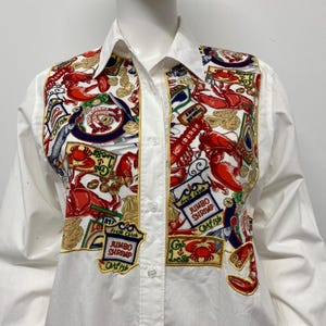 May include: White button-down shirt with a colorful, patterned panel featuring images of seafood, including shrimp, crab, and fish. The panel has gold accents and the text "Jumbo Shrimp" and "Catfish".