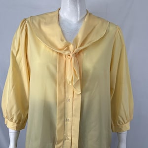 May include: A pale yellow blouse with a rounded collar and a tie detail at the neckline. The long sleeves gather at the cuffs. The blouse has a button-down front and is made of a smooth, lightweight fabric.