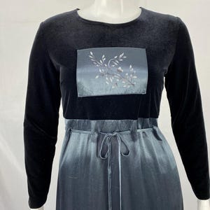 May include: A long-sleeved black velvet dress with a square, satin-like panel on the chest featuring a silver floral design. The dress has a tie-waist and a flowing, ombre-effect skirt in shades of blue and gray.