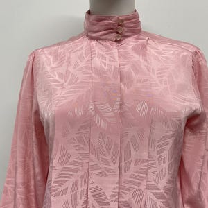 May include: A light pink blouse with a high collar and long sleeves. The fabric has a subtle leaf-like pattern. The collar is fastened with three small buttons. The blouse is made of a silky material.